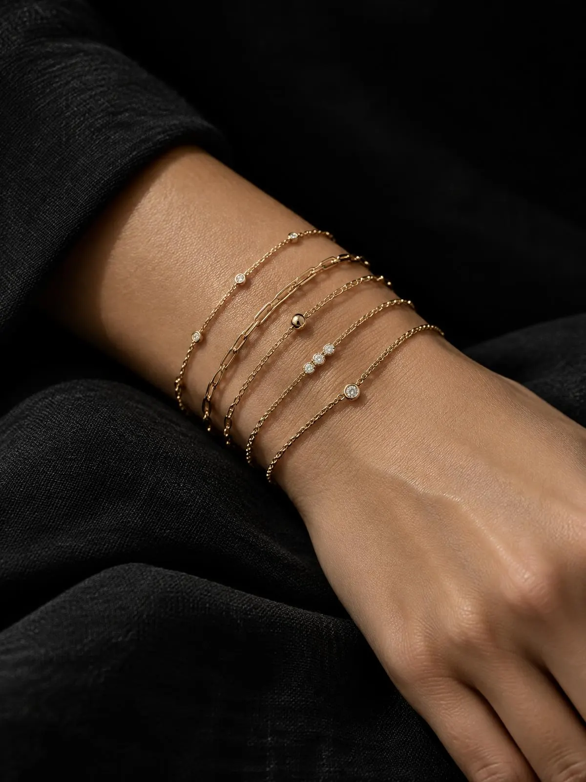 Stack of fine gold chain bracelets on a wrist resting on black linen