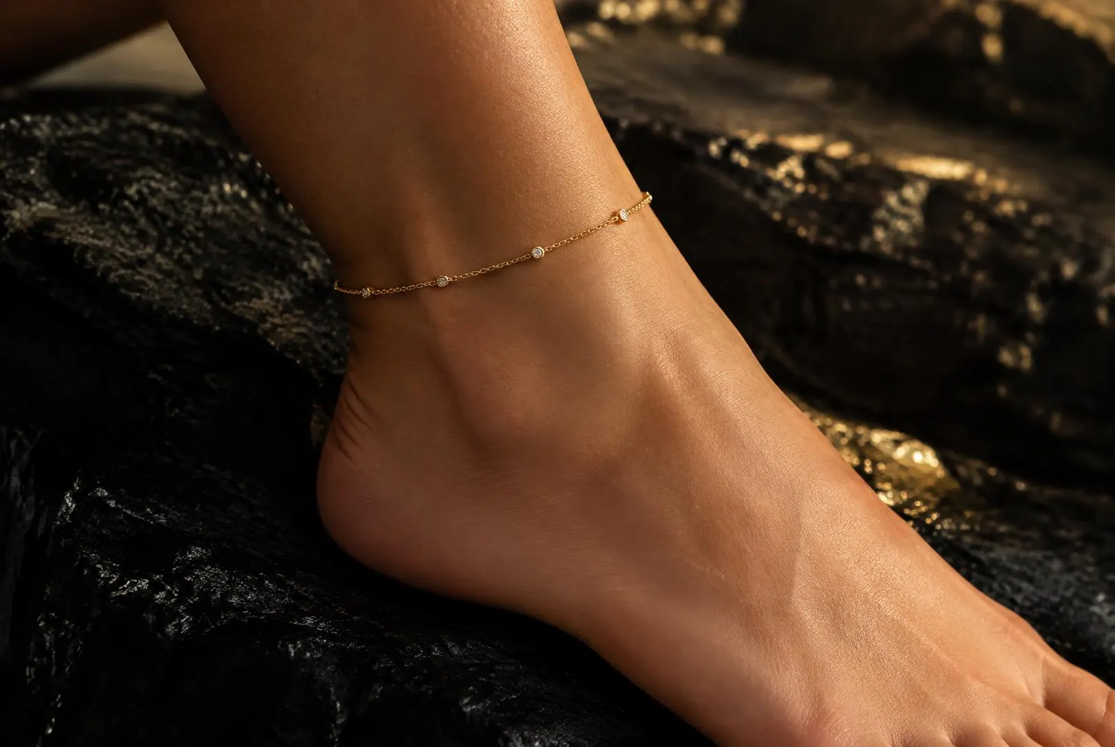 Dainty gold anklet on an ankle against black stone